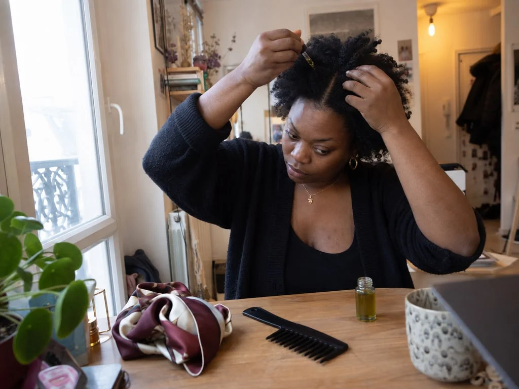 Une femme aux cheveux crépus applique quelques gouttes d'huile sur son cuir chevelu, un foulard en satin posé à côté