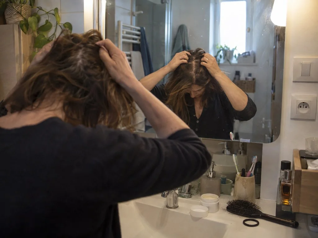 Une femme écarte ses cheveux le long de la raie pour examiner son cuir chevelu dans le miroir d'une salle de bain