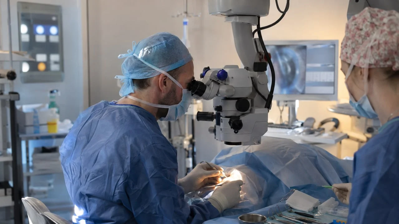 Chirurgien ophtalmologue au microscope opératoire, le visage du patient couvert par un champ stérile